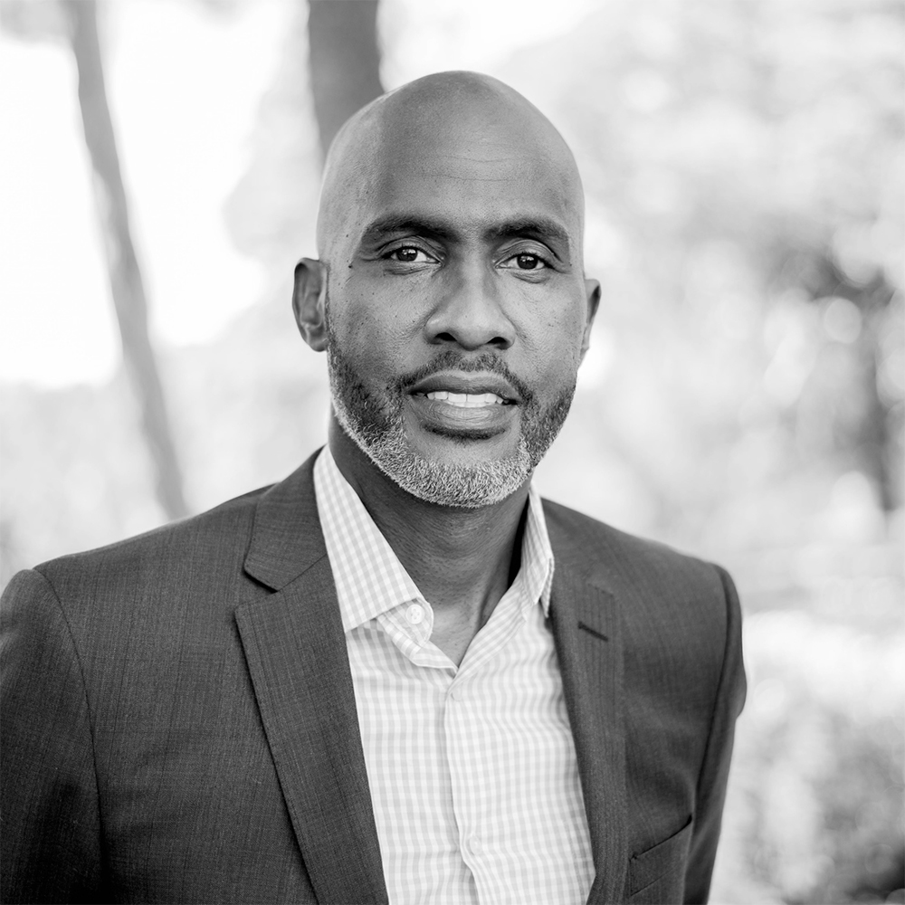 Black and white portrait of Dr. Tyrone C. Howard in a suit standing in front of some foliage