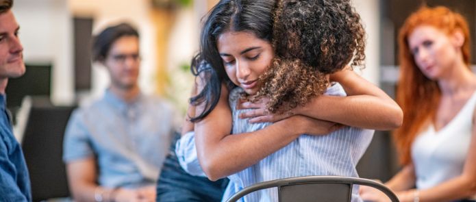 Two women hugging with others sitting down and watching