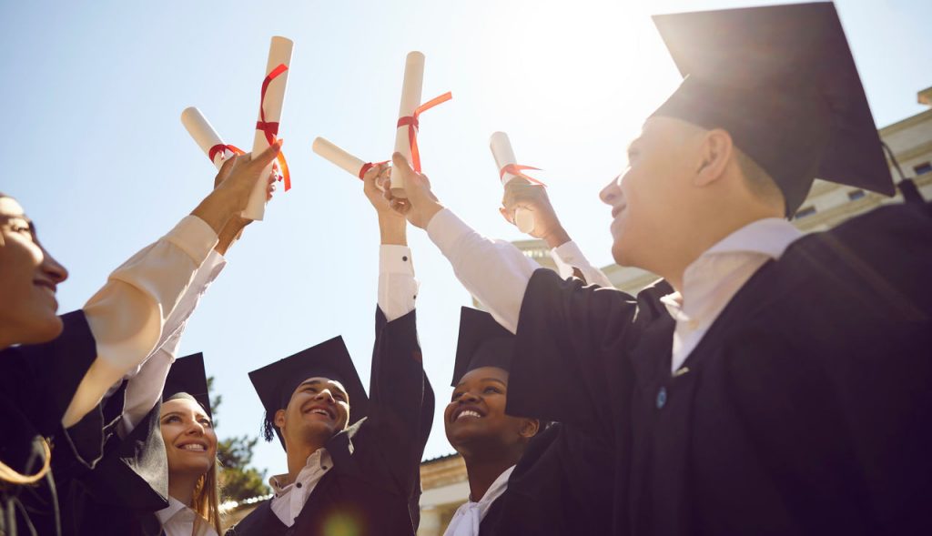 Happy diverse college or university students having fun at graduation. Joyful multiracial graduates standing together and holding up paper diploma scrolls with red ribbons. Education, success