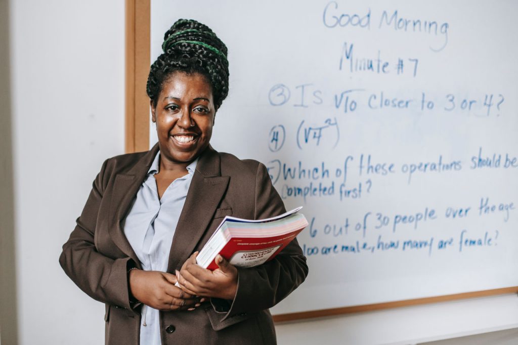 Smiling teacher holding books and standing next to white board