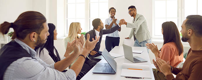 Friendly and diverse employees congratulate man on business achievements, excellent work results or promotion. Business people applaud and cheers during business meeting in bright modern office.