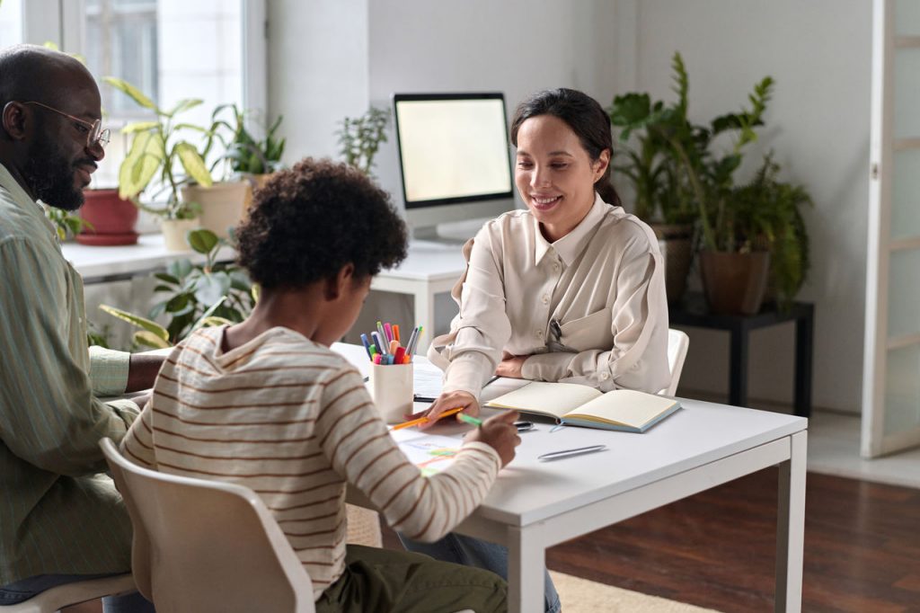 Portrait of smiling young woman as psychologist helping Black family in therapy session with young child drawing pictures